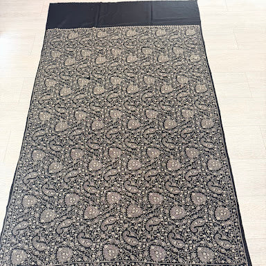 A black and gray patterned shawl or scarf with intricate paisley motifs and a decorative border, laid flat on a light-colored floor.
