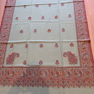A rectangular beige woven textile with red and orange paisley border patterns and small red floral motifs, laid flat on a wooden surface.