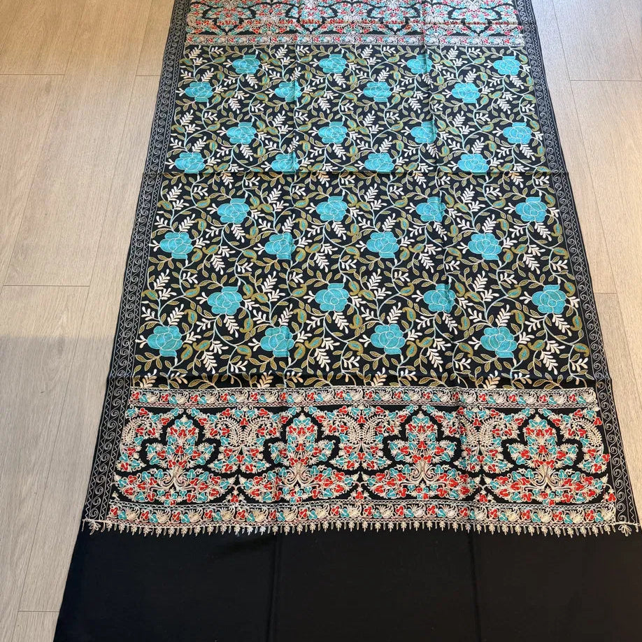 A long black scarf or shawl with intricate floral embroidery, laid flat on a light wood floor. The design features turquoise blue flowers, green leaves, and red accents in the borders.