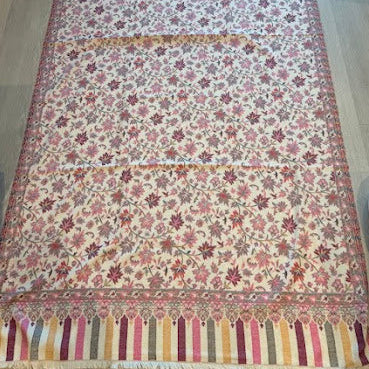 A long rectangular patterned runner rug laid flat on a light wood floor. The rug has an off-white base with a dense floral and vine pattern in pink, mauve, and brown, finished with striped borders in pink, yellow, and brown and fringed ends.