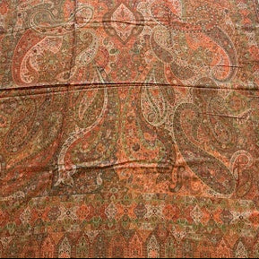 A long rectangular area rug with an intricate paisley and floral pattern in rust orange, brown, and olive green, laid out on a light wood floor between two upholstered chairs.