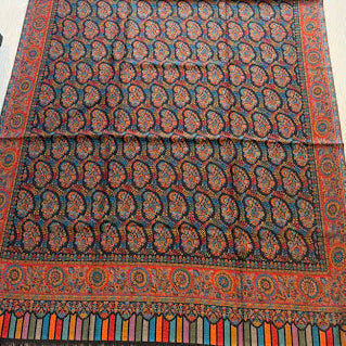A long patterned scarf or shawl laid flat on a light wood floor, featuring a dark navy base with repeating paisley motifs in red, orange, and blue, plus a multicolored striped fringe at the bottom.