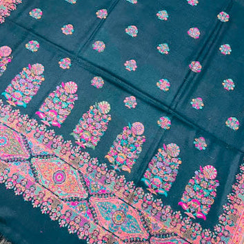 A dark teal black wool shawl with intricate multicolor embroidery, featuring floral motifs and a detailed border, laid flat on a light wood floor.