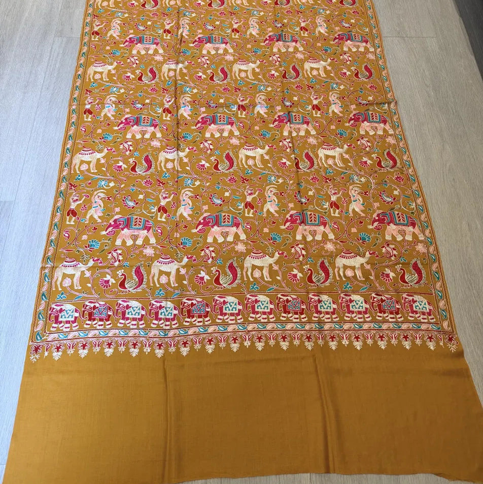 A mustard yellow Banarasi silk saree with intricate multicolor zari embroidery depicting elephants, lions, and floral motifs, laid flat on a light wood floor.