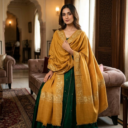 A woman stands in a traditional indoor room wearing a dark green skirt with gold embroidery and a mustard yellow shawl draped over her shoulders. The room features arched doorways, a patterned rug, and upholstered furniture.