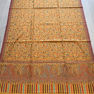 A long patterned textile runner or scarf laid flat on a light wood floor in an indoor room. The fabric has a dense small floral print in yellow, orange, red, and blue, with a decorative border and fringe along one end.