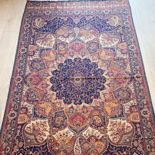A large traditional Persian-style area rug with an intricate floral and medallion pattern, laid out on a light wood floor. The rug features a dominant navy blue base with prominent red, beige, and gold accents in its detailed motifs.