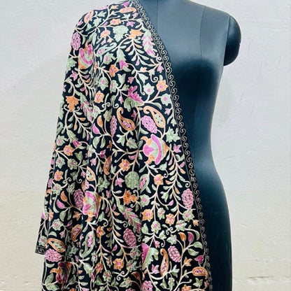 A black embroidered shawl with intricate floral and paisley patterns in pink, green, and orange, draped over a black mannequin torso against a light beige wall.