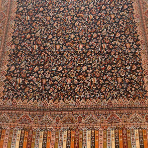 A large traditional oriental rug with an intricate floral pattern, laid out on a light-colored wooden floor. The rug features a dark brown center field surrounded by a warm rust-orange border and detailed fringe at the top and bottom.