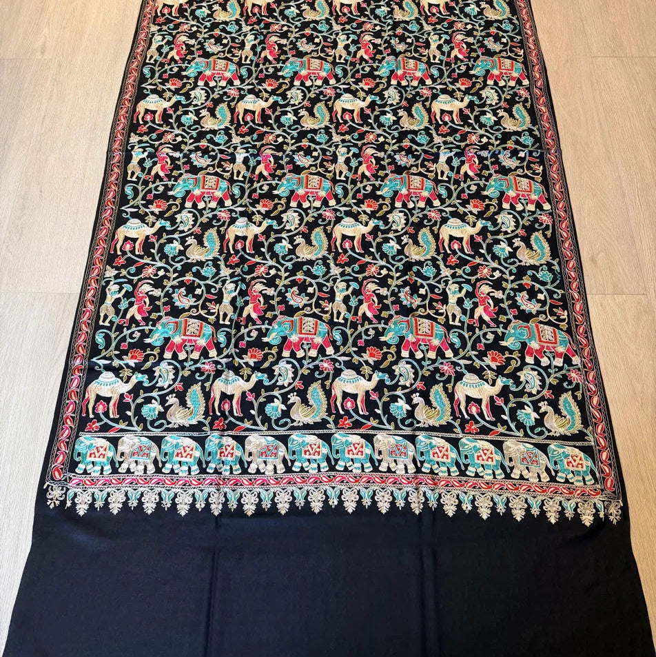 A black embroidered textile, likely a shawl or scarf, featuring intricate multicolor hand-embroidery with elephants, deer, and floral motifs on a light wood floor background.