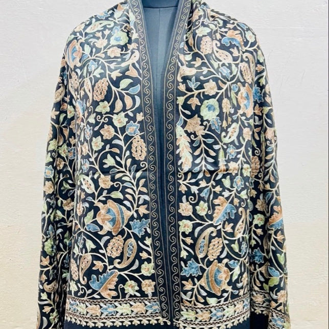 A dark navy blue embroidered shawl or jacket with intricate floral and paisley patterns in beige, light blue, and green, displayed on a mannequin against a light textured wall.