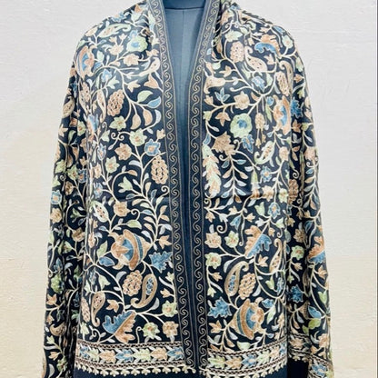 A dark navy blue embroidered shawl or jacket with intricate floral and paisley patterns in beige, light blue, and green, displayed on a mannequin against a light textured wall.