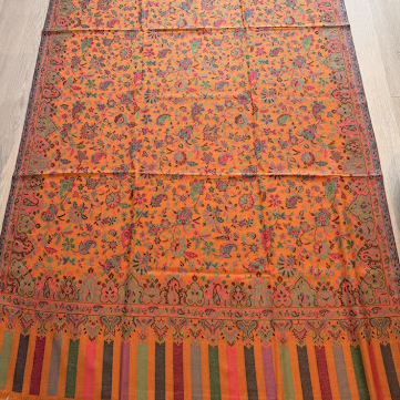 A long patterned textile laid flat on a light wood floor, featuring an orange base with dense multicolored floral motifs and striped borders in red, green, blue, and yellow.