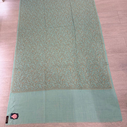 A sage green and beige patterned shawl or scarf laid flat on a light wood floor, featuring a solid sage green border and a small embroidered tag near the bottom left.