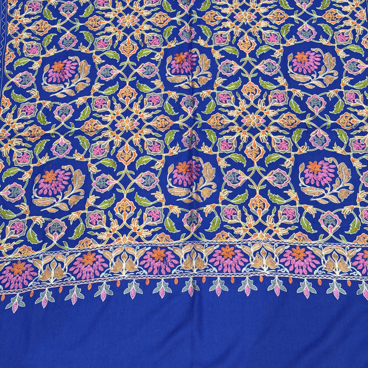 A close-up of a richly patterned royal blue fabric, likely a silk saree or textile, featuring intricate floral and geometric motifs in pink, purple, green, and gold.