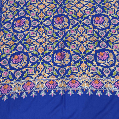 A close-up of a richly patterned royal blue fabric, likely a silk saree or textile, featuring intricate floral and geometric motifs in pink, purple, green, and gold.