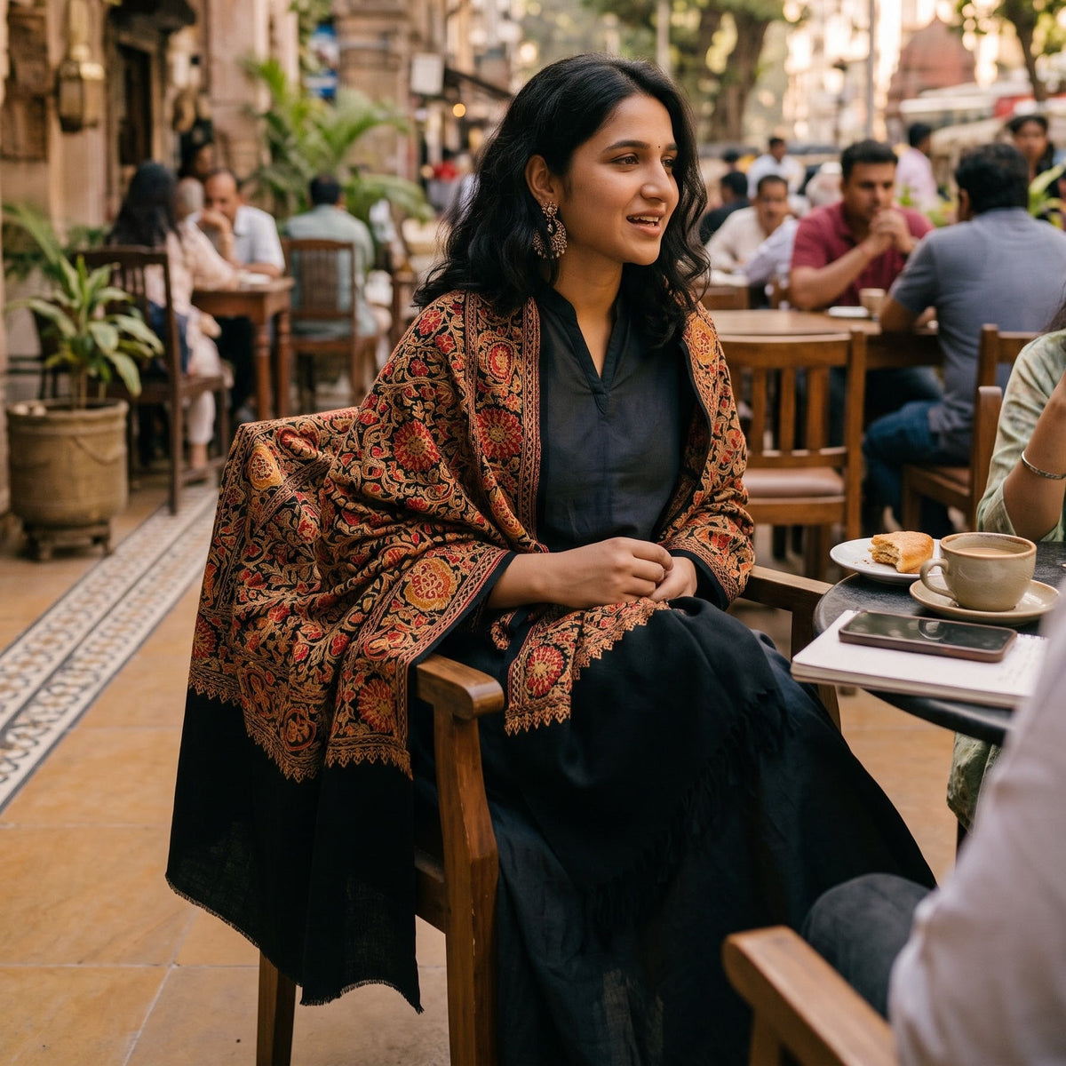 A woman seated at an outdoor cafe table wearing a black outfit and a rust-orange patterned shawl with a black border. The background shows a warm, bustling street scene with other patrons and buildings.