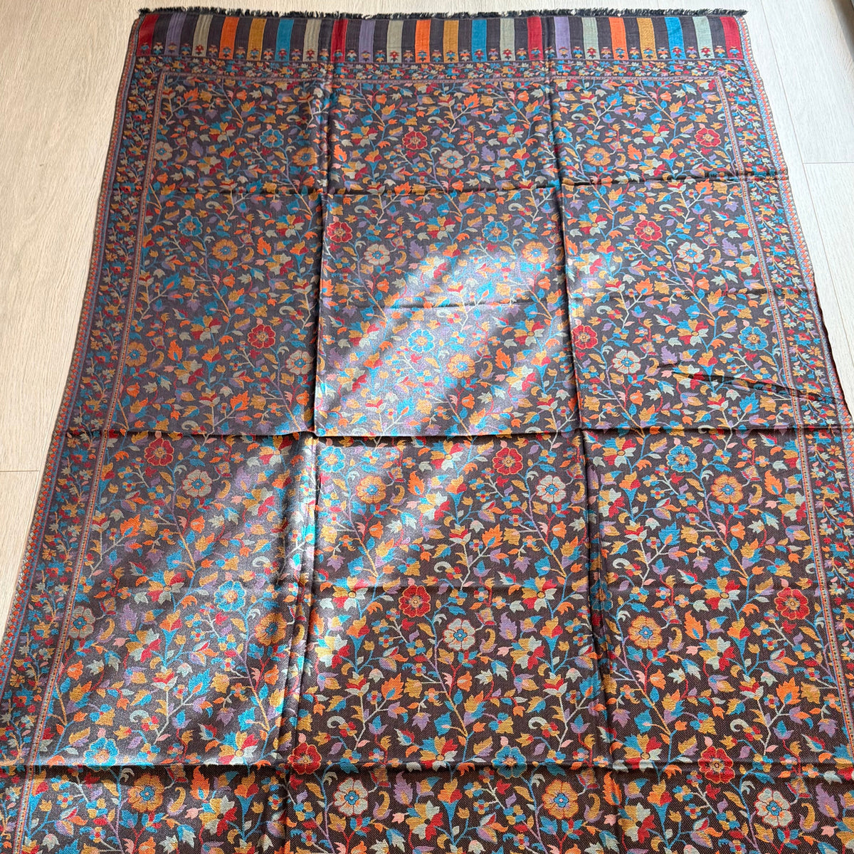 A large rectangular patterned textile laid flat on a light wood floor, featuring an intricate floral and geometric print with multicolored stripes along the borders and fringe at the bottom. A rolled gray fabric or rug is partially visible on the right side.