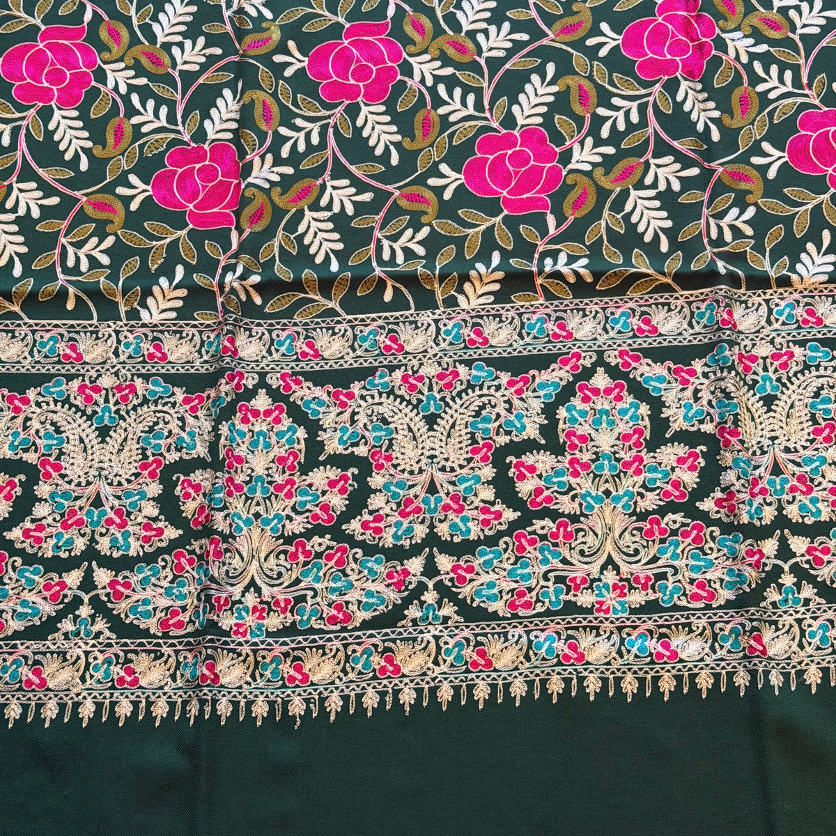 A close-up of a patterned textile with a dark charcoal base, bright pink floral motifs, white leafy vines, and an ornate multicolored border featuring turquoise, pink, beige, and black details.