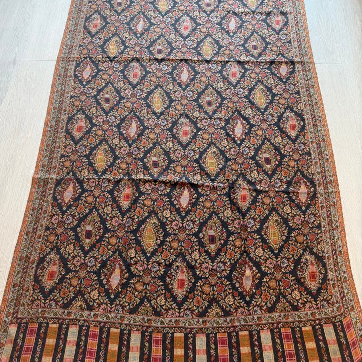 A long patterned area rug laid flat on a light wood floor, featuring a dark navy base with repeating diamond motifs in rust red, beige, and orange, plus a striped border at the ends.