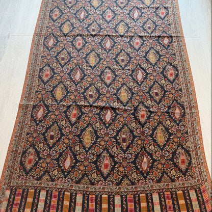 A long patterned area rug laid flat on a light wood floor, featuring a dark navy base with repeating diamond motifs in rust red, beige, and orange, plus a striped border at the ends.