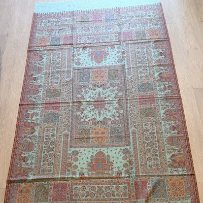 A rectangular patterned shawl or scarf laid flat on a light wood floor, featuring a detailed traditional design in muted sage green, terracotta, rust, and cream tones.