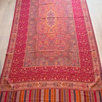 A traditional Persian-style area rug laid out on a light wood floor, featuring a vibrant red and orange floral pattern with striped fringe at the ends.