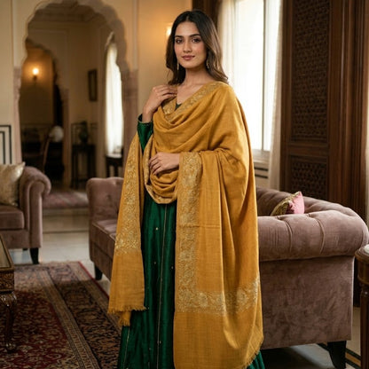 A woman models a traditional Indian outfit featuring a deep green skirt, a mustard yellow shawl draped over her shoulders, and a matching mustard yellow dupatta with gold embroidery, set in an ornate, traditional room with arches and sofas.
