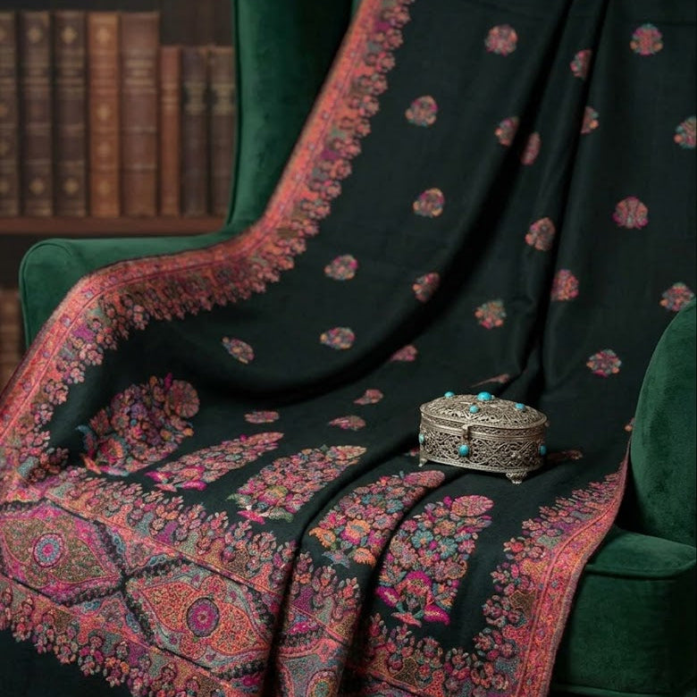 A dark green shawl with intricate pink, red, and gold floral patterns and a wide border, draped over a green velvet armchair next to a silver decorative box.