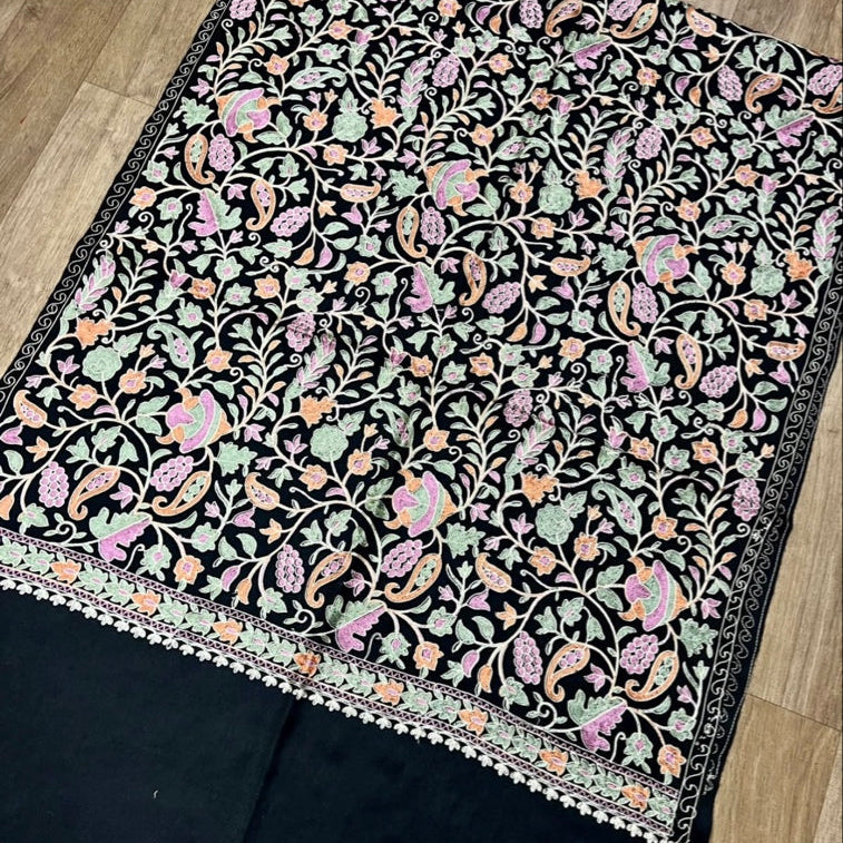 A black embroidered fabric piece, likely a dupatta or scarf, featuring intricate floral and paisley patterns in pink, green, and orange, laid flat on a light wood floor.