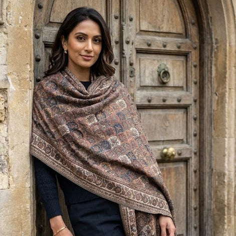 A woman wearing a brown patterned shawl draped over her shoulders, standing in front of a large rustic wooden door.