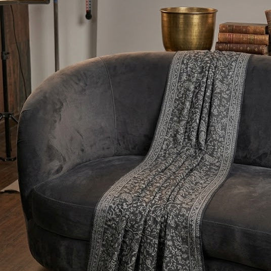 A dark gray patterned throw blanket with fringe is draped over a dark gray velvet sofa in a room with wooden floors and a brass bowl in the background.