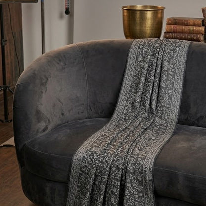 A dark gray patterned throw blanket with fringe is draped over a dark gray velvet sofa in a room with wooden floors and a brass bowl in the background.