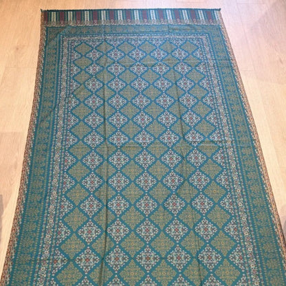 A long rectangular patterned runner rug laid flat on a light wood floor, with a teal and green geometric design and striped fringe along one end.