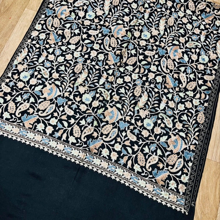 A black shawl with an intricate floral and paisley pattern in beige, light blue, and white, laid flat on a light wood floor.