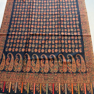A long black woven textile with dense orange and beige floral motifs and a large paisley border, laid flat on a light wood floor.