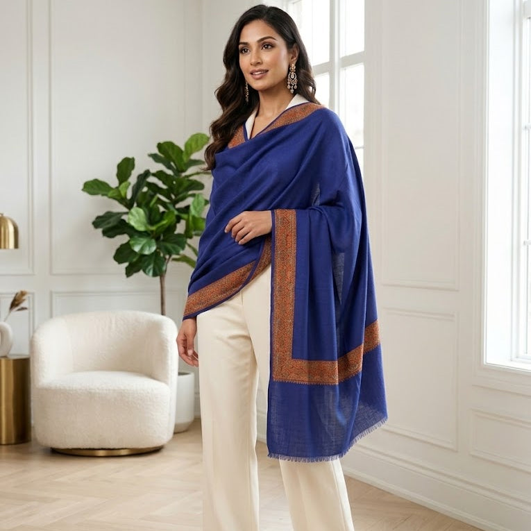 A woman models a royal blue shawl with brown border details, paired with cream wide-leg trousers, in a bright, modern room with white walls and a wooden floor.