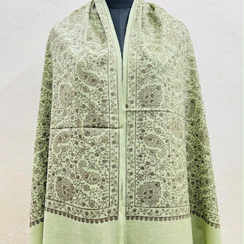 A light green shawl with intricate beige and brown floral embroidery, displayed on a black mannequin torso against a plain off-white wall.