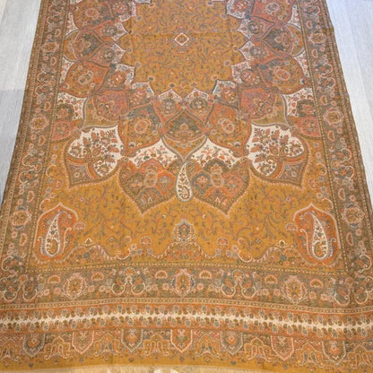A rectangular traditional area rug with an intricate floral and geometric pattern, laid out on a light-colored wooden floor. The dominant color is a warm ochre yellow, complemented by brown, beige, and muted red accents in the detailed pattern.