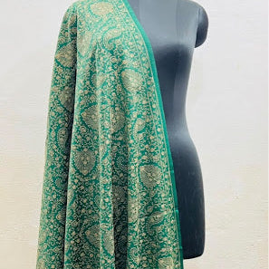 A green shawl with intricate gold floral patterns draped over a black mannequin torso against a light beige wall.