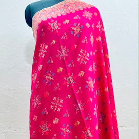 A bright pink traditional draped fabric, likely a dupatta or sari, featuring intricate multicolor floral and geometric block prints, displayed on a black mannequin torso against a white wall.