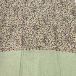 A light sage green and beige patterned shawl or scarf laid flat on a light wood floor. The fabric features a dense, all-over floral and paisley print in muted brown and beige tones, with solid sage green borders at the top and bottom.