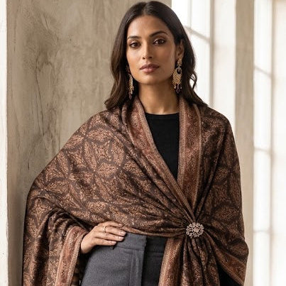 A woman models a brown and beige patterned shawl wrap with a decorative brooch, styled over a black top and charcoal gray trousers in a sunlit indoor setting.