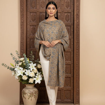 Woman in a patterned top and white pants standing next to a decorative vase with flowers, against a beige wall.