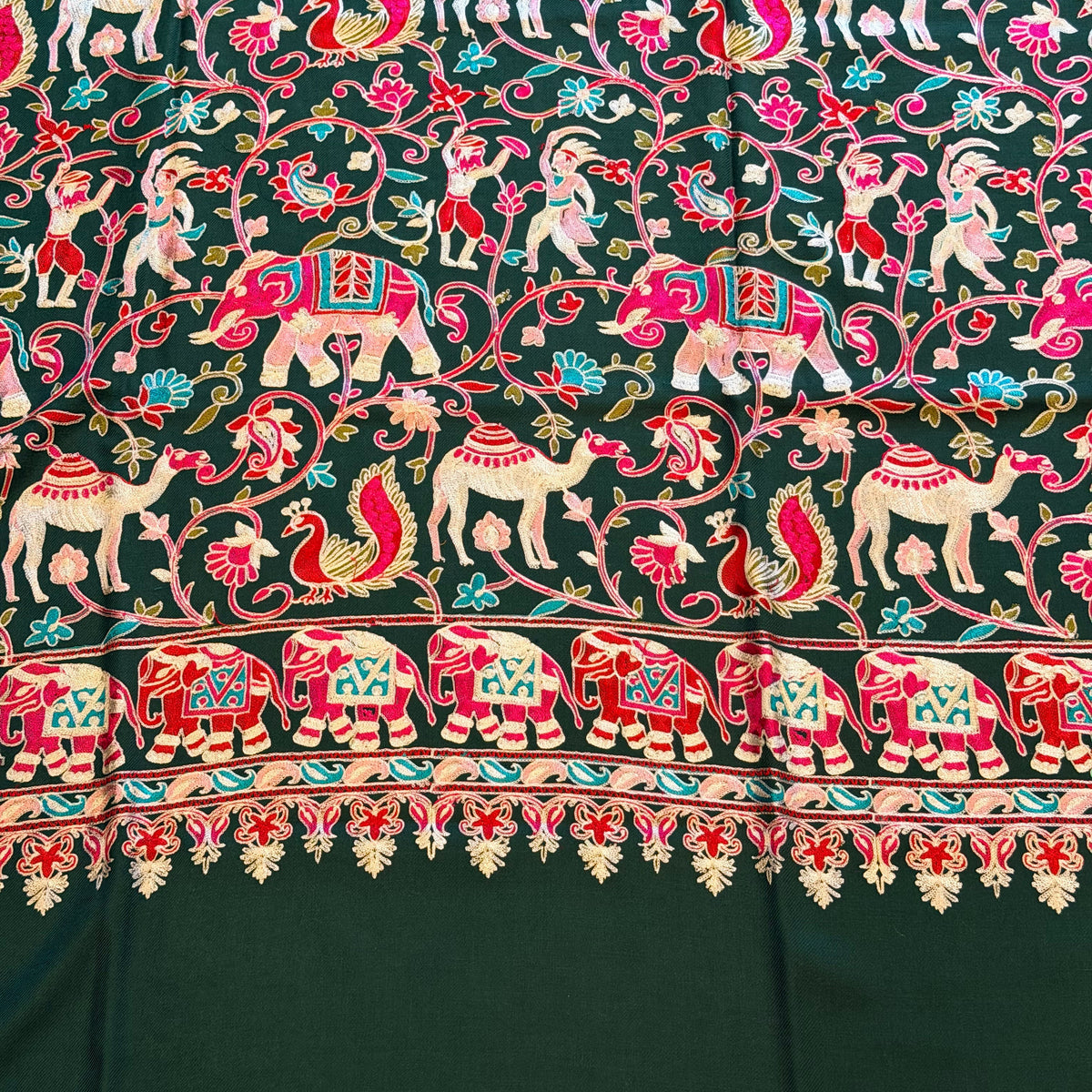 A close-up of a dark green fabric with an intricate hand-block printed pattern featuring pink, red, white, and light blue elephants, floral vines, and a decorative border.
