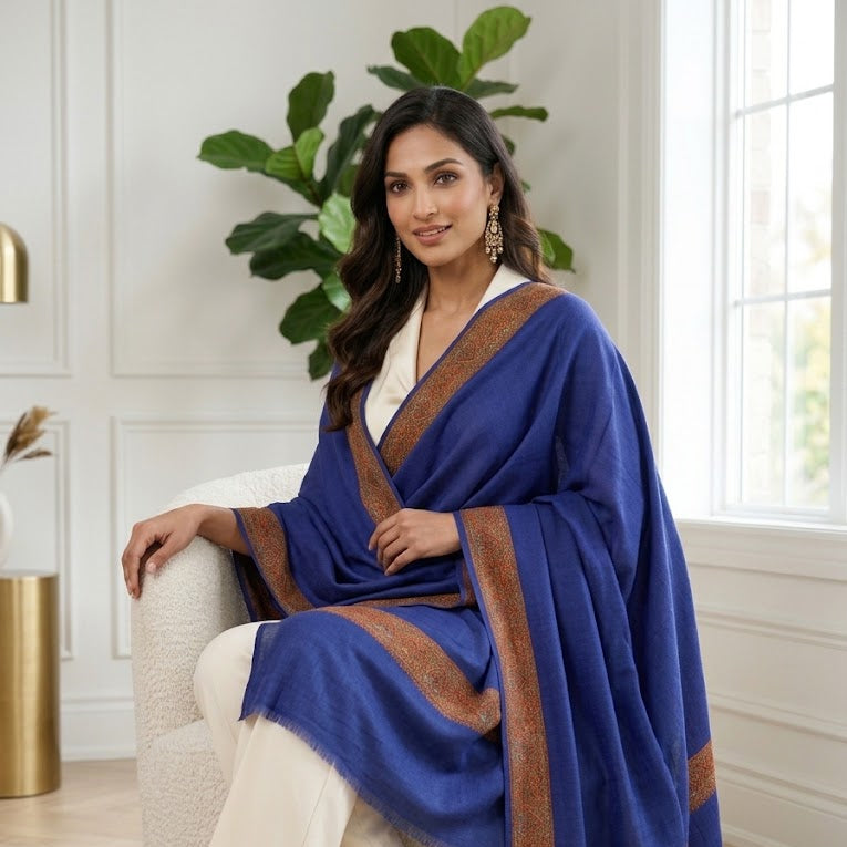 A woman sits on a white armchair in a bright room, wearing a royal blue shawl with brown border details over a white outfit.