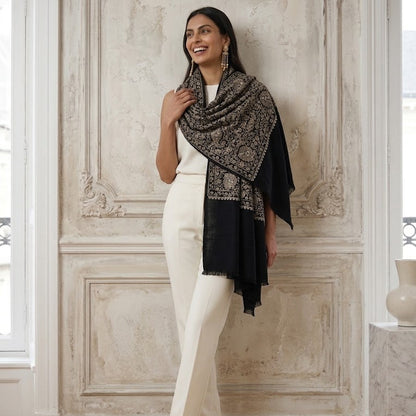 A woman stands in a classical interior wearing a black and beige patterned shawl over a white outfit.