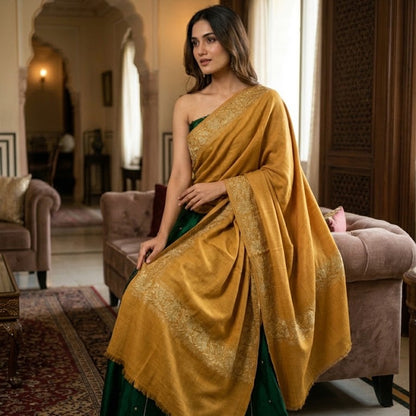 A woman wearing a mustard yellow and green traditional Indian saree with gold embroidery, standing in a decorated room with traditional arches and sofas.