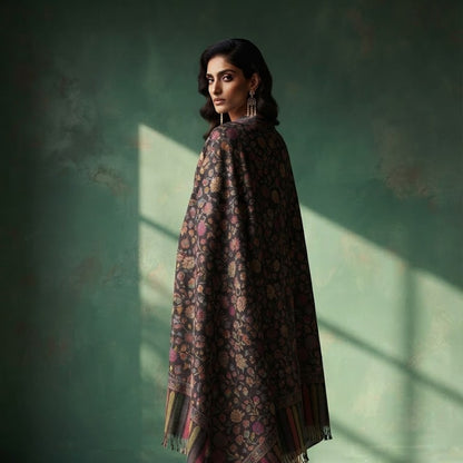 A woman stands in profile wearing a dark patterned shawl draped over her shoulders, paired with white pants and beige heels, against a textured green wall with soft window light casting shadows.