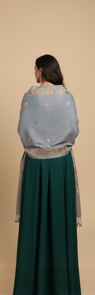 Woman wearing a traditional outfit with a light blue embroidered shawl and green skirt on a beige background